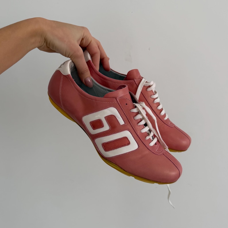May include: A pair of pink leather sneakers with white laces and the number "60" in white on the side. The soles are yellow, and the heel and trim are white. The shoes are held up by a hand.