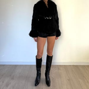 Peut inclure: Veste noire en fausse fourrure avec un large col et une ceinture &agrave; boucle argent&eacute;e. La veste a des manches longues avec des poignets &eacute;vas&eacute;s. Associ&eacute;e &agrave; un short noir et des bottes en cuir noires montantes. L'ensemble est pos&eacute; contre un mur blanc et un sol en bois clair.