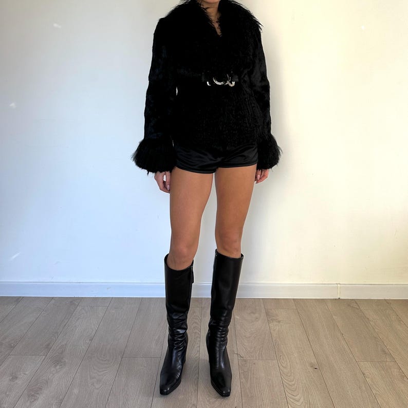 Peut inclure: Veste noire en fausse fourrure avec un large col et une ceinture &agrave; boucle argent&eacute;e. La veste a des manches longues avec des poignets &eacute;vas&eacute;s. Associ&eacute;e &agrave; un short noir et des bottes en cuir noires montantes. L'ensemble est pos&eacute; contre un mur blanc et un sol en bois clair.