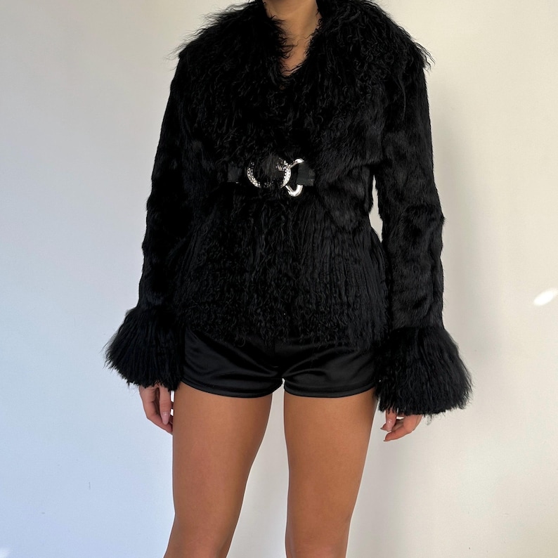 Peut inclure: Veste en fausse fourrure noire avec un large col et des manches &eacute;vas&eacute;es. La veste est cintr&eacute;e &agrave; la taille par une ceinture noire avec une boucle argent&eacute;e. La texture de la veste est hirsute, et le style g&eacute;n&eacute;ral est chic.