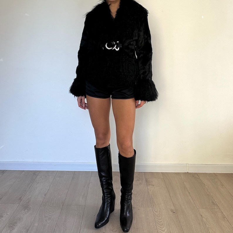 Peut inclure: Veste noire en fausse fourrure avec une large ceinture et des manches &eacute;vas&eacute;es. La veste est associ&eacute;e &agrave; un short noir et &agrave; des bottes hautes en cuir noir. La tenue est pr&eacute;sent&eacute;e devant un mur blanc uni et un sol en bois clair.