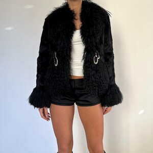 Peut inclure: Un manteau en fausse fourrure noire avec une texture particuli&egrave;re. Le manteau a un large col, des manches &eacute;vas&eacute;es et des fermoirs argent&eacute;s. Le mod&egrave;le porte un haut blanc et un short noir. La longueur du manteau est d'environ 70 cm.