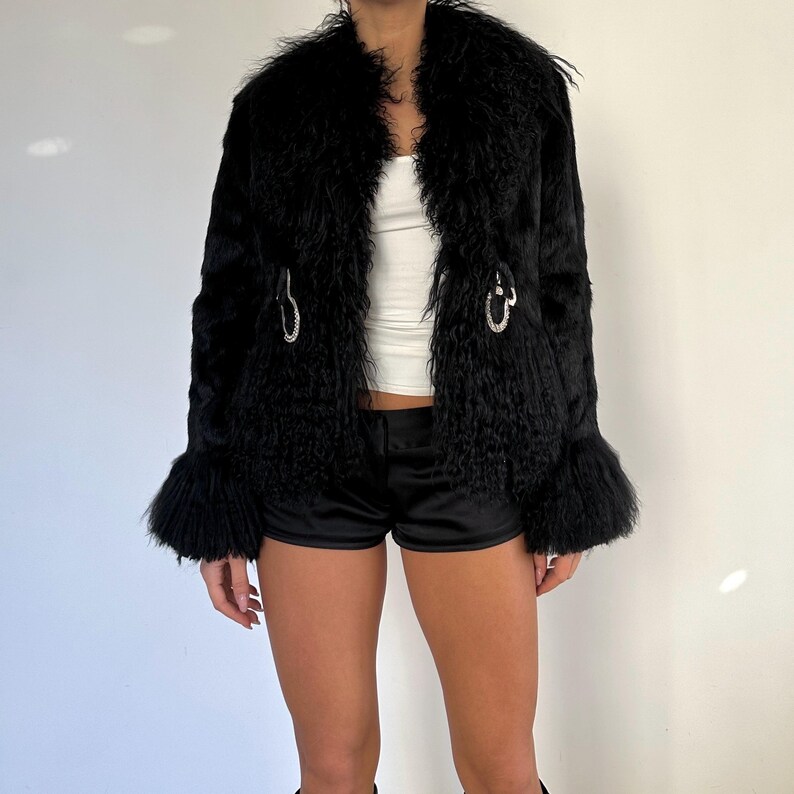 Peut inclure: Un manteau en fausse fourrure noire avec une texture particuli&egrave;re. Le manteau a un large col, des manches &eacute;vas&eacute;es et des fermoirs argent&eacute;s. Le mod&egrave;le porte un haut blanc et un short noir. La longueur du manteau est d'environ 70 cm.