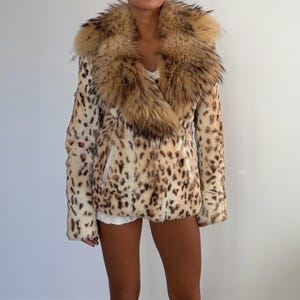 May include: A hip-length jacket featuring a cream-colored base with a brown leopard print pattern. The garment includes a large, voluminous, fluffy brown and tan faux fur collar that extends down the front, long sleeves, and a straight hemline.