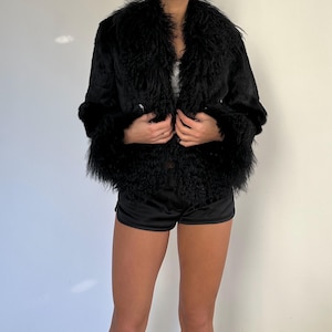 Peut inclure: Veste en fausse fourrure noire avec un large col et des poignets. La veste est ouverte, r&eacute;v&eacute;lant un haut blanc en dessous. Le mod&egrave;le porte &eacute;galement un short noir. La veste a un aspect textur&eacute;.