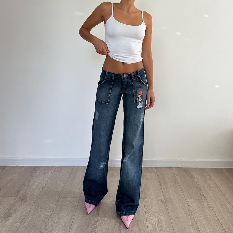 May include: Dark wash denim jeans with a low-rise waist and wide legs. The jeans feature distressed details and a tag. The model is wearing a white tank top and pink heels. A casual, stylish outfit.