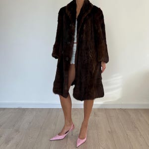 May include: A long, dark brown fur coat with a button-up front and a wide collar. The coat is knee-length and has long sleeves. The person is wearing pink heels.