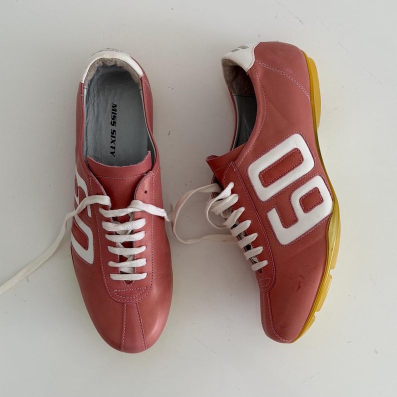 May include: Pair of pink leather sneakers with white laces and accents. The sneakers feature the number "60" in white on the side and the text "MISS SIXTY" on the insole. The soles are yellow.