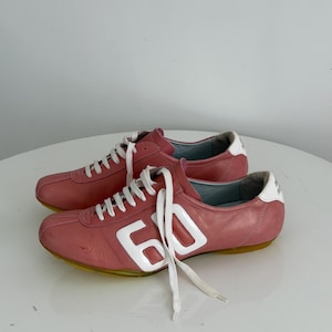 May include: A pair of pink leather sneakers with white laces and the number "60" in white on the side. The sneakers have a white heel accent and a yellow sole. The shoes are on a white surface.