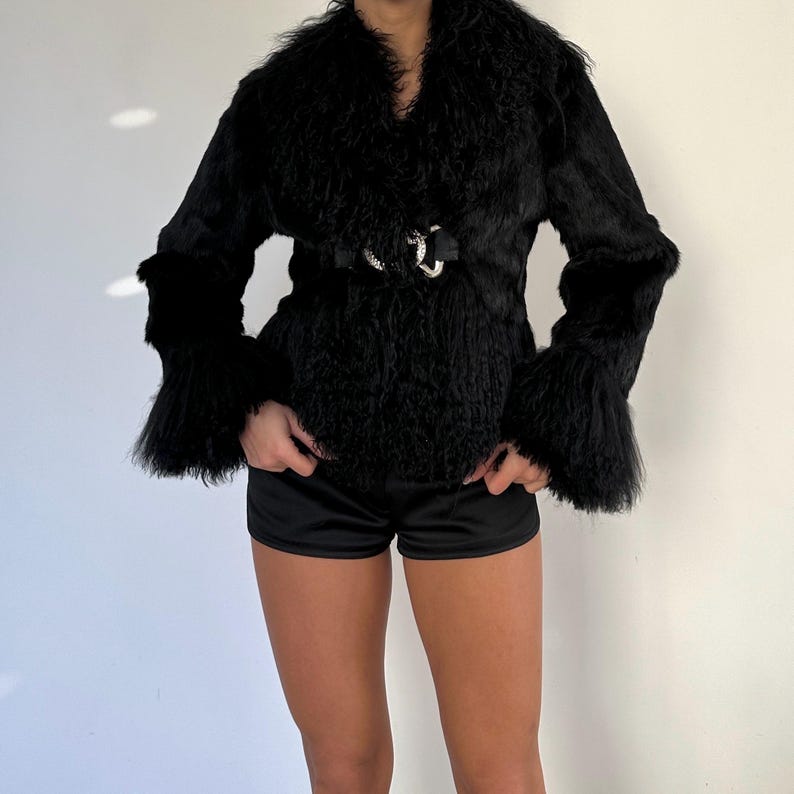 Peut inclure: Veste en fausse fourrure noire avec un large col et des manches &eacute;vas&eacute;es. La veste est cintr&eacute;e &agrave; la taille avec une ceinture noire et une boucle argent&eacute;e. V&ecirc;tement d'ext&eacute;rieur &eacute;l&eacute;gant.