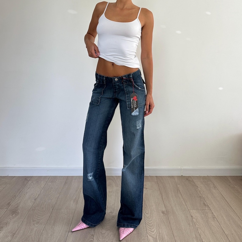 May include: Dark wash denim jeans with a low rise and wide leg. The jeans have distressed details and a tag attached to the front pocket. The model is wearing a white tank top and pink pointed-toe heels.