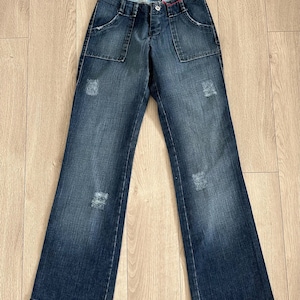 May include: Dark wash denim jeans with a flared leg, featuring front pockets and distressed details. The jeans are laid flat on a wooden surface, showcasing their design and style. A classic casual wear item.