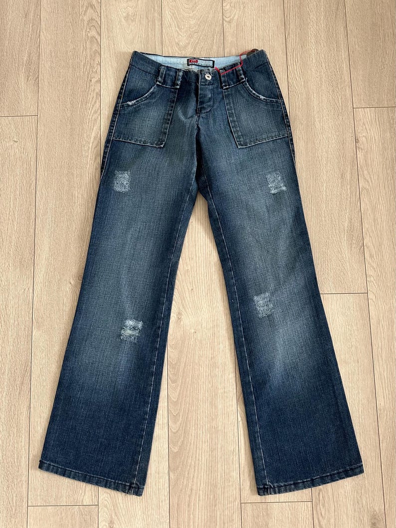 May include: Dark wash denim jeans with a flared leg, featuring front pockets and distressed details. The jeans are laid flat on a wooden surface, showcasing their design and style. A classic casual wear item.