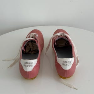 May include: A pair of pink sneakers with white laces. The sneakers have a white patch on the heel with the words "MISS SIXTY" in silver lettering. The soles are yellow.
