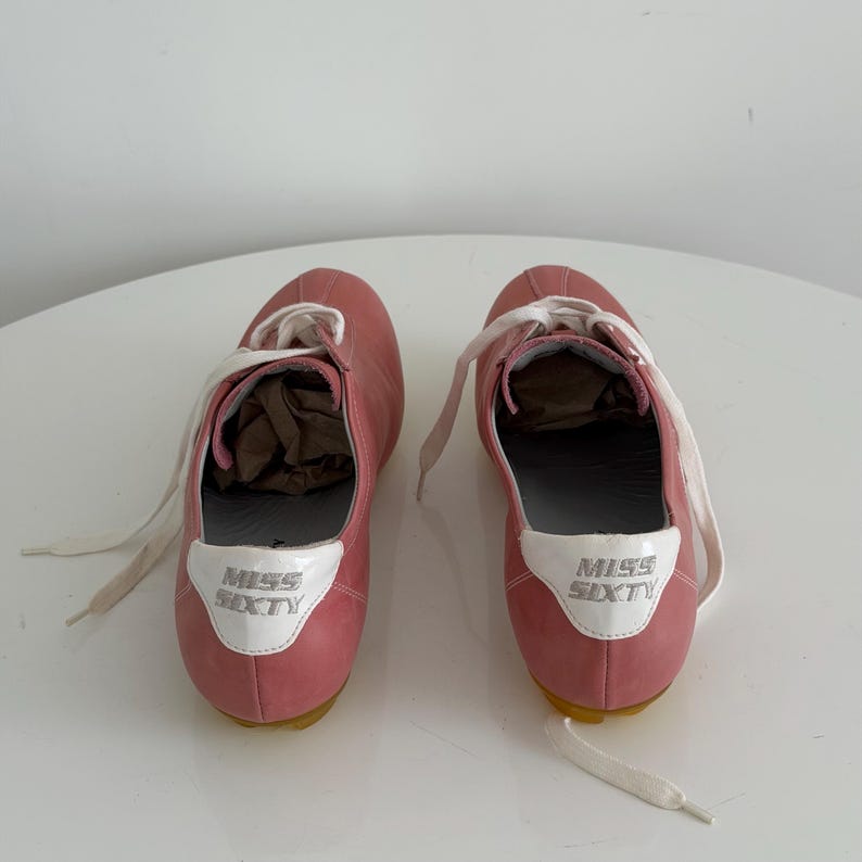 May include: A pair of pink sneakers with white laces. The sneakers have a white patch on the heel with the words "MISS SIXTY" in silver lettering. The soles are yellow.