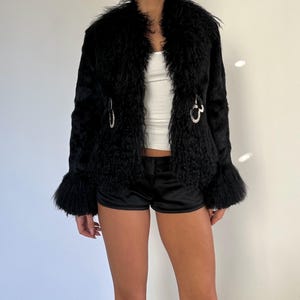 Peut inclure: Veste en fausse fourrure noire &agrave; l'aspect textur&eacute; et hirsute. La veste pr&eacute;sente un large col, des manches &eacute;vas&eacute;es et des fermoirs d&eacute;coratifs argent&eacute;s. L'ensemble est compl&eacute;t&eacute; par un short noir et un haut blanc.