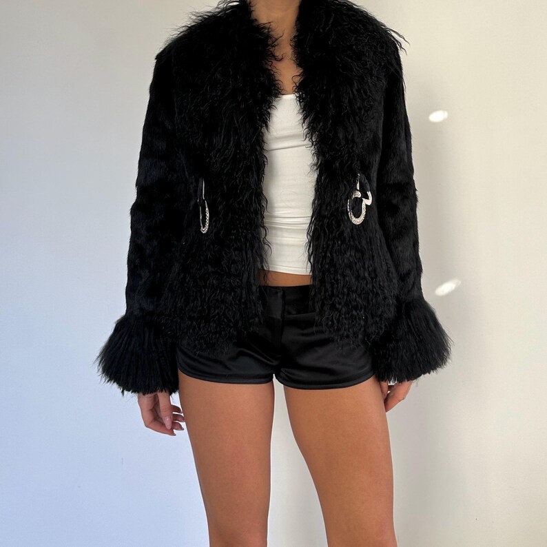 Peut inclure: Veste en fausse fourrure noire &agrave; l'aspect textur&eacute; et hirsute. La veste pr&eacute;sente un large col, des manches &eacute;vas&eacute;es et des fermoirs d&eacute;coratifs argent&eacute;s. L'ensemble est compl&eacute;t&eacute; par un short noir et un haut blanc.