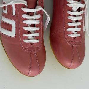 May include: Pair of dusty rose sneakers with white accents and laces. The sneakers have a retro design with a white stripe on the side and a yellow sole. The shoes are made of leather.