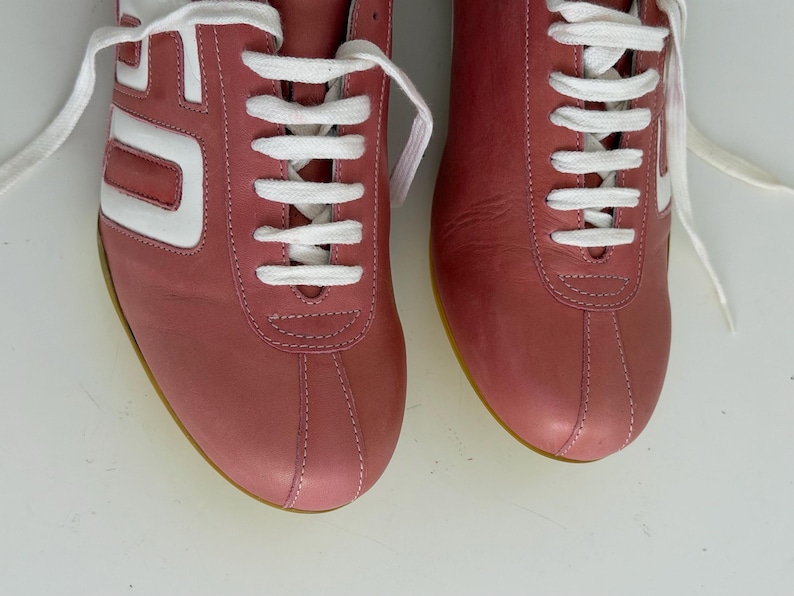 May include: Pair of dusty rose sneakers with white accents and laces. The sneakers have a retro design with a white stripe on the side and a yellow sole. The shoes are made of leather.