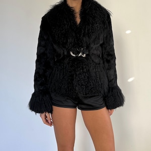 Peut inclure: Veste en fausse fourrure noire avec un large col et une ceinture d&eacute;corative &agrave; boucle argent&eacute;e. La veste a des manches longues avec des poignets &eacute;vas&eacute;s. La veste est port&eacute;e avec un short noir.