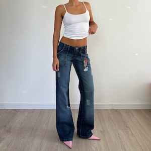 May include: Dark wash denim jeans with a wide leg and distressed details. The jeans have a low-rise waist, front pockets, and a button closure. A white tank top is worn with the jeans. The model is wearing pink pointed-toe shoes.