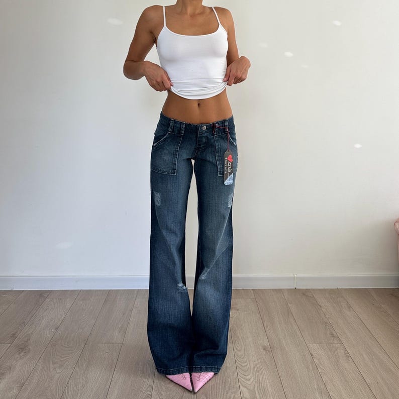 May include: Dark wash denim jeans with a wide leg design. The jeans have a low-rise waist, front pockets, and distressed details. A white tank top is worn with the jeans. Pink pointed-toe shoes complete the outfit.