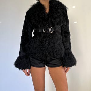 Peut inclure: Veste en fausse fourrure noire avec un large col et des manches &eacute;vas&eacute;es. La veste est cintr&eacute;e &agrave; la taille par une ceinture noire avec une boucle argent&eacute;e. La veste est associ&eacute;e &agrave; un short noir.