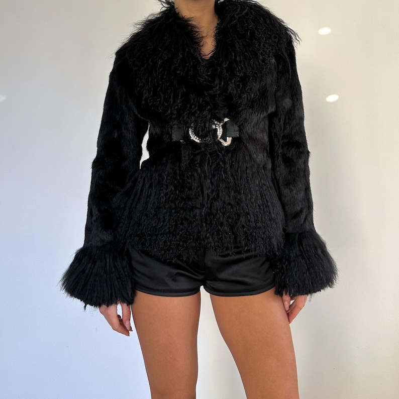 Peut inclure: Veste en fausse fourrure noire avec un large col et des manches &eacute;vas&eacute;es. La veste est cintr&eacute;e &agrave; la taille par une ceinture noire avec une boucle argent&eacute;e. La veste est associ&eacute;e &agrave; un short noir.