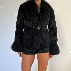 Peut inclure: Un manteau en fausse fourrure noire avec un large col et des manches &eacute;vas&eacute;es. La taille est cintr&eacute;e par une ceinture noire avec une boucle argent&eacute;e. La texture du manteau est hirsute, et le style g&eacute;n&eacute;ral est vintage.