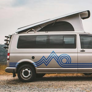 May include: A silver camper van with a pop-up roof parked on a gravel track near the coast. The van features blue wave graphics and a rear rack. The sky is blue with clouds.