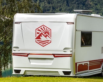 May include: A white travel trailer with red accents and a mountain logo on the back. The trailer is parked on a grassy area with a lake and mountains in the background. The image is taken during the day.