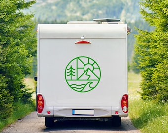 May include: A white recreational vehicle, or RV, drives down a dirt road lined with tall green trees. The back of the RV features a green circular graphic with a mountain, sun, tree, and wave design. A snow-capped mountain is visible in the background.