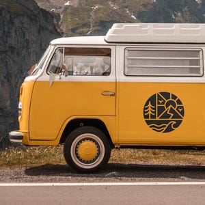 May include: A yellow and white vintage camper van with a black circular graphic on the side, parked on a road with a mountain backdrop. The graphic features trees, a sun, waves, and mountains. The van has a white roof.