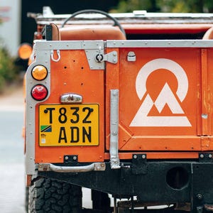 May include: Close-up of an orange vintage SUV with a white mountain logo. The vehicle has a yellow license plate with black lettering and a green and white emblem. The rear lights are visible.