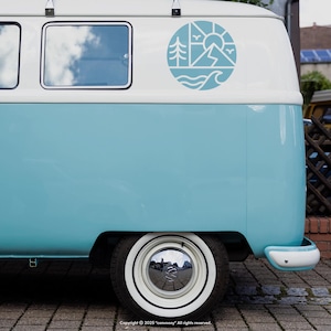 May include: A light blue and white vintage van with a circular decal featuring a mountain, sun, trees, and waves. The van has a white wheel with a chrome hubcap and a white bumper. The van is parked on a brick surface.