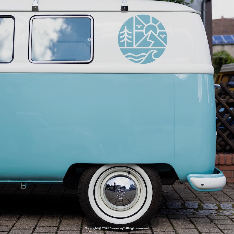 May include: A light blue and white vintage van with a circular decal featuring a mountain, sun, trees, and waves. The van has a white wheel with a chrome hubcap and a white bumper. The van is parked on a brick surface.