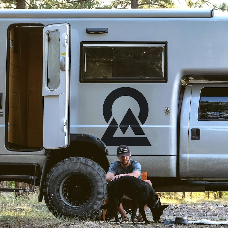 Großer schwarzer Berg-Emblem-Aufkleber im Kreis auf grauem Expeditionsmobil; Mann streichelt Hund am Waldcamp – CONVOOOY Vinylsticker.
