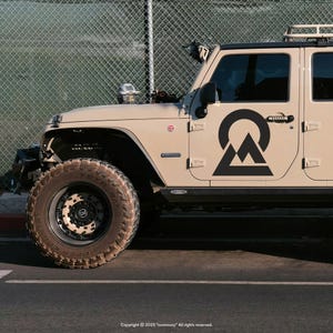 Großer schwarzer Berg-Emblem Vinylaufkleber auf der Fahrertür eines beigen Jeep Wrangler Offroad-Fahrzeugs vor Maschendrahtzaun, CONVOOOY Design.