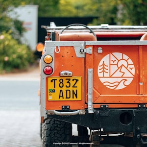 May include: Close-up of the rear of an orange vintage off-road vehicle. The vehicle has a yellow license plate with black text and a white graphic of a mountain, sun, trees, and waves. The vehicle's taillights are visible.