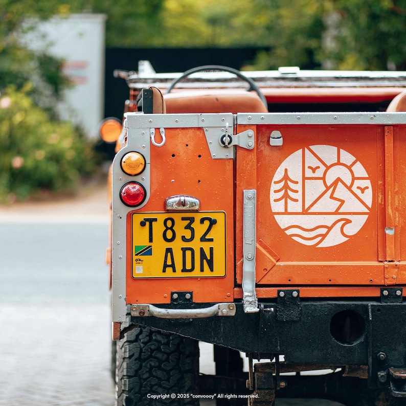May include: Close-up of the rear of an orange vintage off-road vehicle. The vehicle has a yellow license plate with black text and a white graphic of a mountain, sun, trees, and waves. The vehicle's taillights are visible.