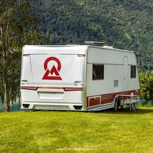 Großer schwarzer Berg-Emblem Vinylaufkleber auf der Heckklappe eines weißen Wohnwagens, geparkt auf grüner Wiese vor See- und Berglandschaft.