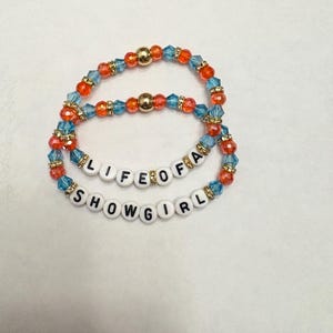 Puede incluir: Dos pulseras de cuentas. La pulsera superior tiene cuentas naranjas, azules y doradas. La pulsera inferior tiene el texto "LIFE OF A SHOWGIRL" escrito con cuentas de letras blancas. Las pulseras est&aacute;n sobre un fondo blanco.