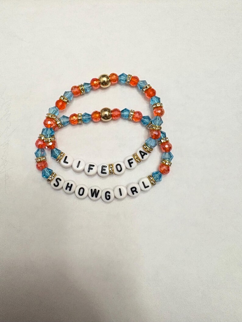 Puede incluir: Dos pulseras de cuentas. La pulsera superior tiene cuentas naranjas, azules y doradas. La pulsera inferior tiene el texto "LIFE OF A SHOWGIRL" escrito con cuentas de letras blancas. Las pulseras est&aacute;n sobre un fondo blanco.