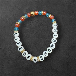 May include: A bracelet with white letter beads spelling "LIFE OF A SHOW GIRL". The bracelet features orange, blue, and gold-coloured beads. The bracelet is on a dark grey surface.