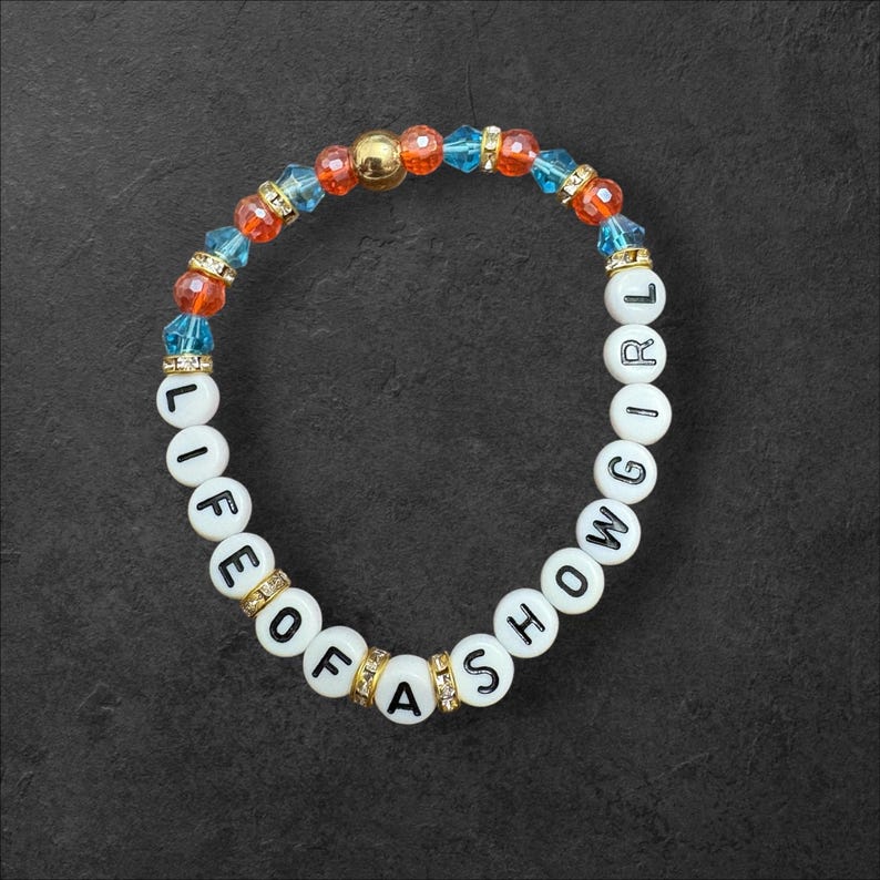 May include: A bracelet with white letter beads spelling "LIFE OF A SHOW GIRL". The bracelet features orange, blue, and gold-coloured beads. The bracelet is on a dark grey surface.