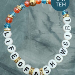 May include: A beaded bracelet with white letter beads spelling "LIFE OF A SHOWGIRL". The bracelet features orange, blue, and gold beads, with gold and rhinestone accents. A blue circle with "NEW ITEM" is in the background.