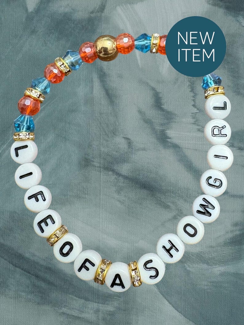 May include: A beaded bracelet with white letter beads spelling "LIFE OF A SHOWGIRL". The bracelet features orange, blue, and gold beads, with gold and rhinestone accents. A blue circle with "NEW ITEM" is in the background.