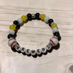 May include: A beaded bracelet featuring yellow, navy blue, and white beads, two baseball-shaped beads, and white letter beads spelling BREWERS. The design includes silver-toned spacers and clear rhinestone-encrusted accents between the beads.