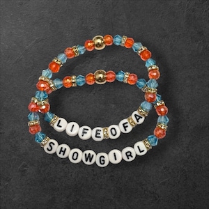 Puede incluir: Dos pulseras de cuentas con las palabras "LIFE OF A SHOWGIRL". Las pulseras presentan cuentas naranjas, azules y doradas. Las cuentas est&aacute;n ensartadas en una goma el&aacute;stica. Las pulseras est&aacute;n sobre un fondo gris oscuro.