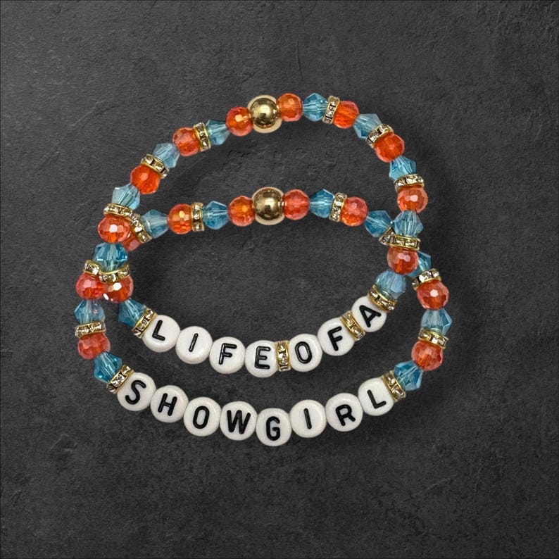 Puede incluir: Dos pulseras de cuentas con las palabras "LIFE OF A SHOWGIRL". Las pulseras presentan cuentas naranjas, azules y doradas. Las cuentas est&aacute;n ensartadas en una goma el&aacute;stica. Las pulseras est&aacute;n sobre un fondo gris oscuro.